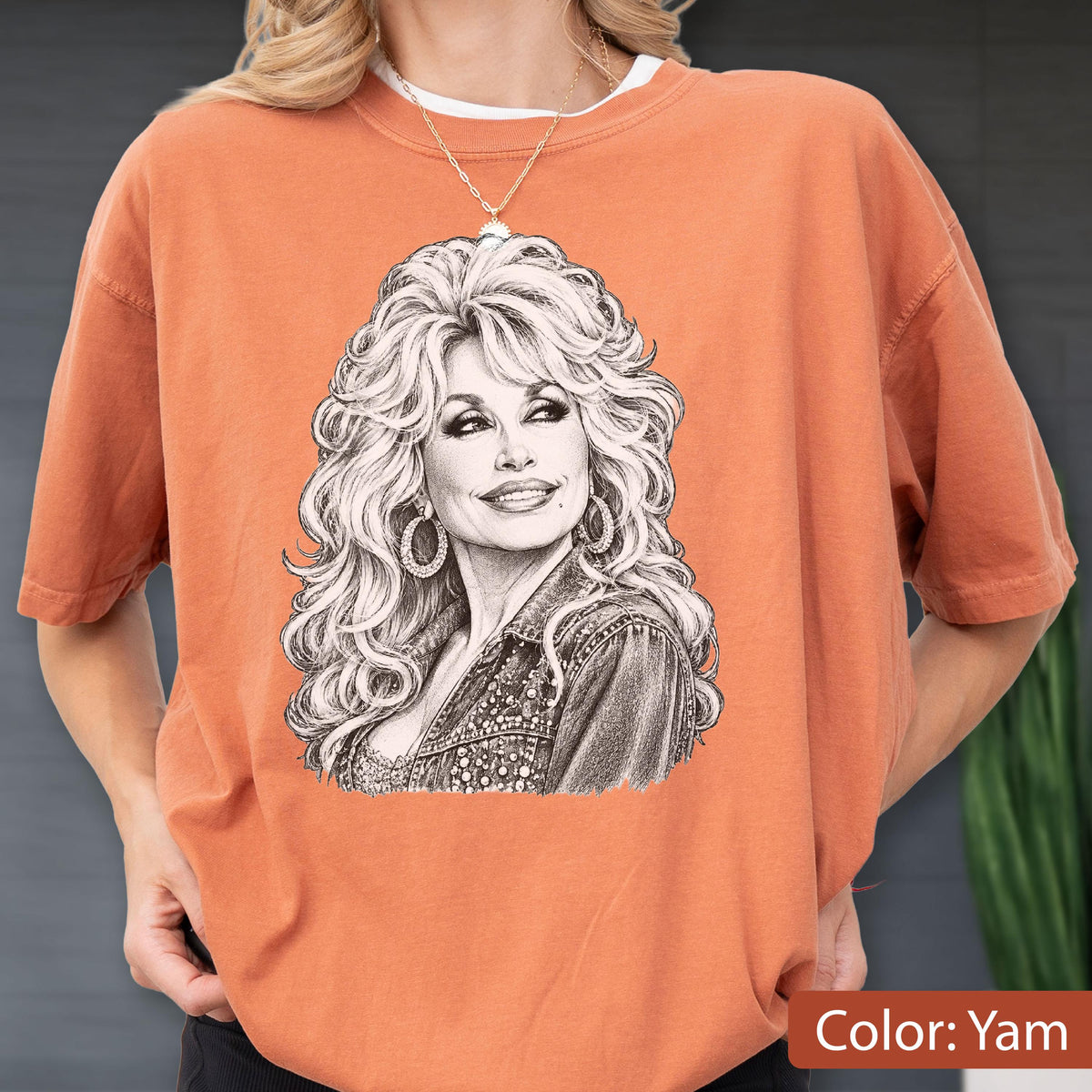 May include: A burnt orange crewneck t-shirt featuring a detailed black and white sketch of a woman with voluminous blonde hair and large hoop earrings. The graphic is centered on the chest, showcasing a portrait of the individual wearing a jacket.