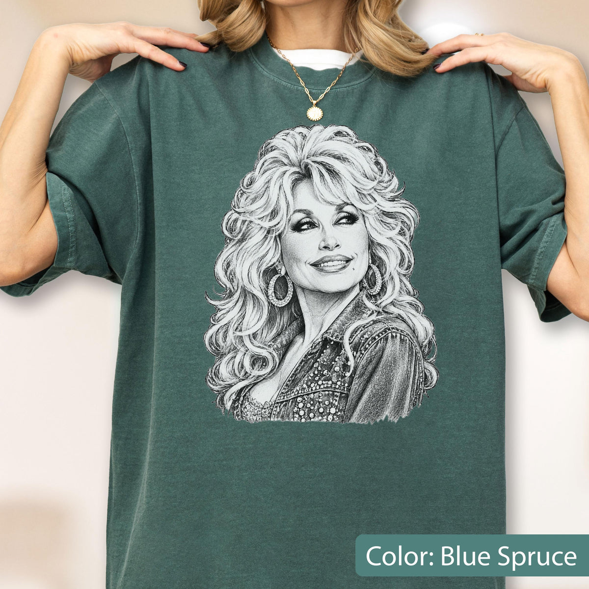 May include: A blue spruce short-sleeve t-shirt featuring a detailed black and white sketch graphic of a woman with voluminous blonde hair and large hoop earrings. The illustration depicts a portrait of a smiling individual wearing a collared jacket.