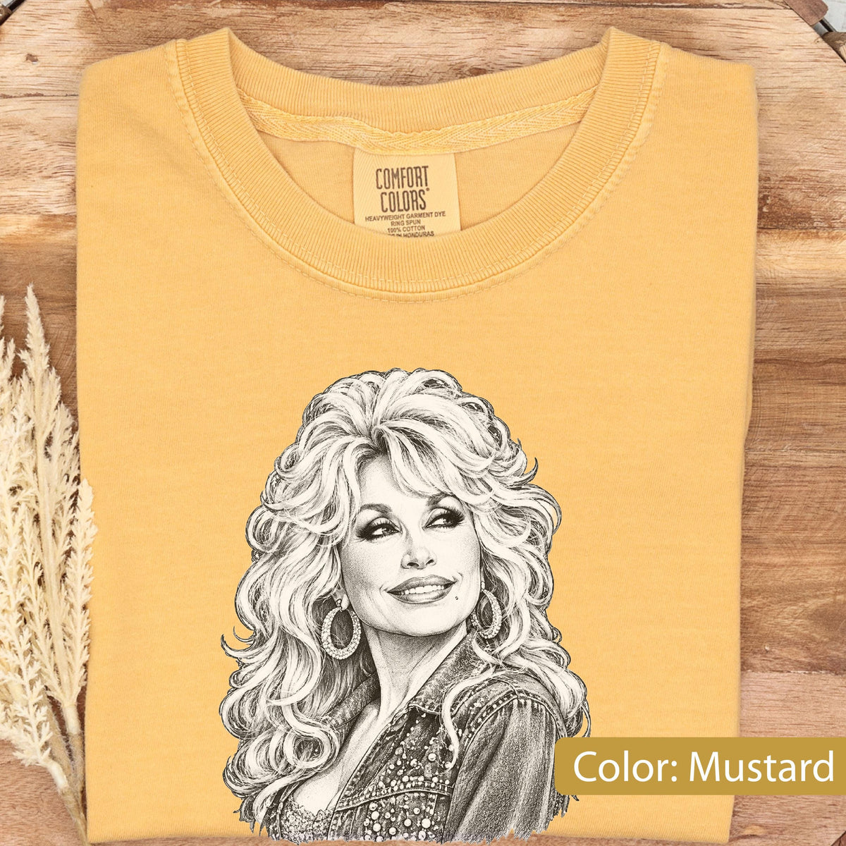 May include: A mustard yellow t-shirt featuring a black and white sketch of a person with voluminous blonde hair, wearing hoop earrings and a textured jacket. The inner neck label displays the text Comfort Colors, Heavyweight Garment Dye.