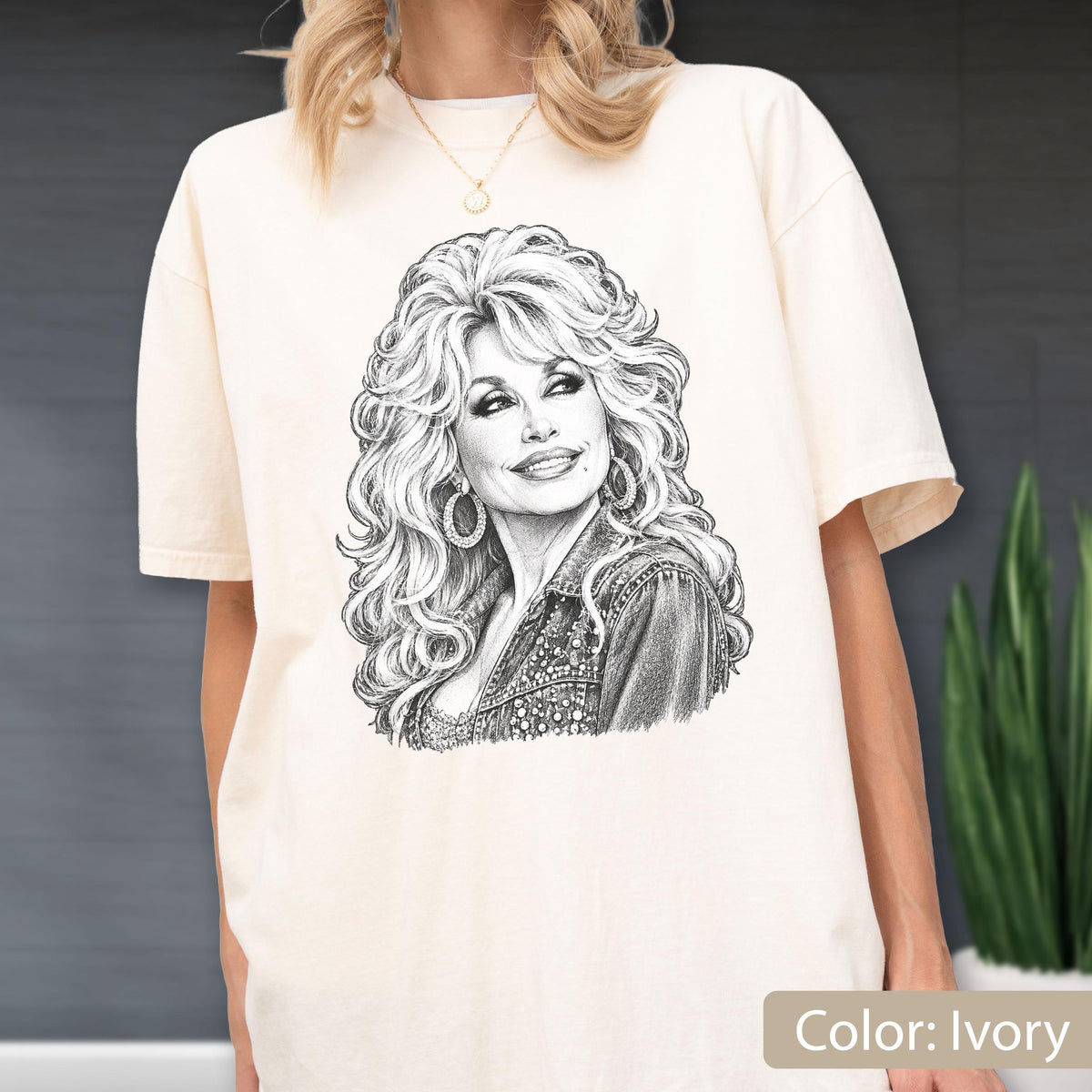 May include: An ivory short-sleeve t-shirt featuring a detailed black and white pencil-style sketch of a woman with voluminous blonde hair, wearing hoop earrings and a textured jacket, centered on the front of the garment.