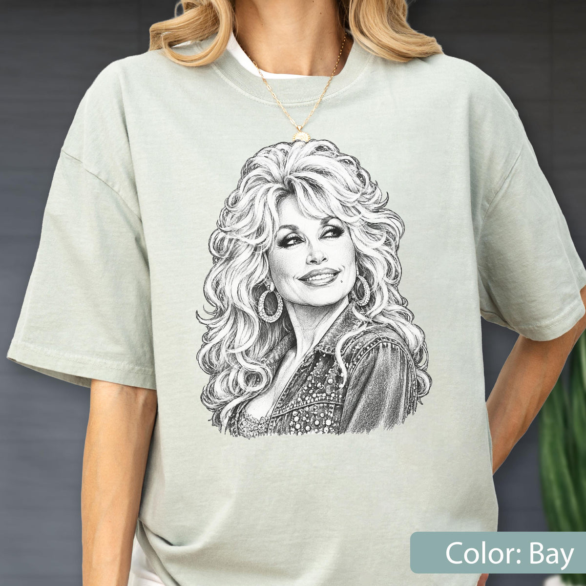 May include: A light sage green short-sleeve t-shirt featuring a detailed black and white sketch of a woman with voluminous blonde hair, wearing large hoop earrings and a textured jacket. The graphic is centered on the chest of the garment.