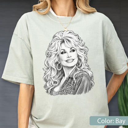 May include: A light sage green short-sleeve t-shirt featuring a detailed black and white sketch of a woman with voluminous blonde hair, wearing large hoop earrings and a textured jacket. The graphic is centered on the chest of the garment.