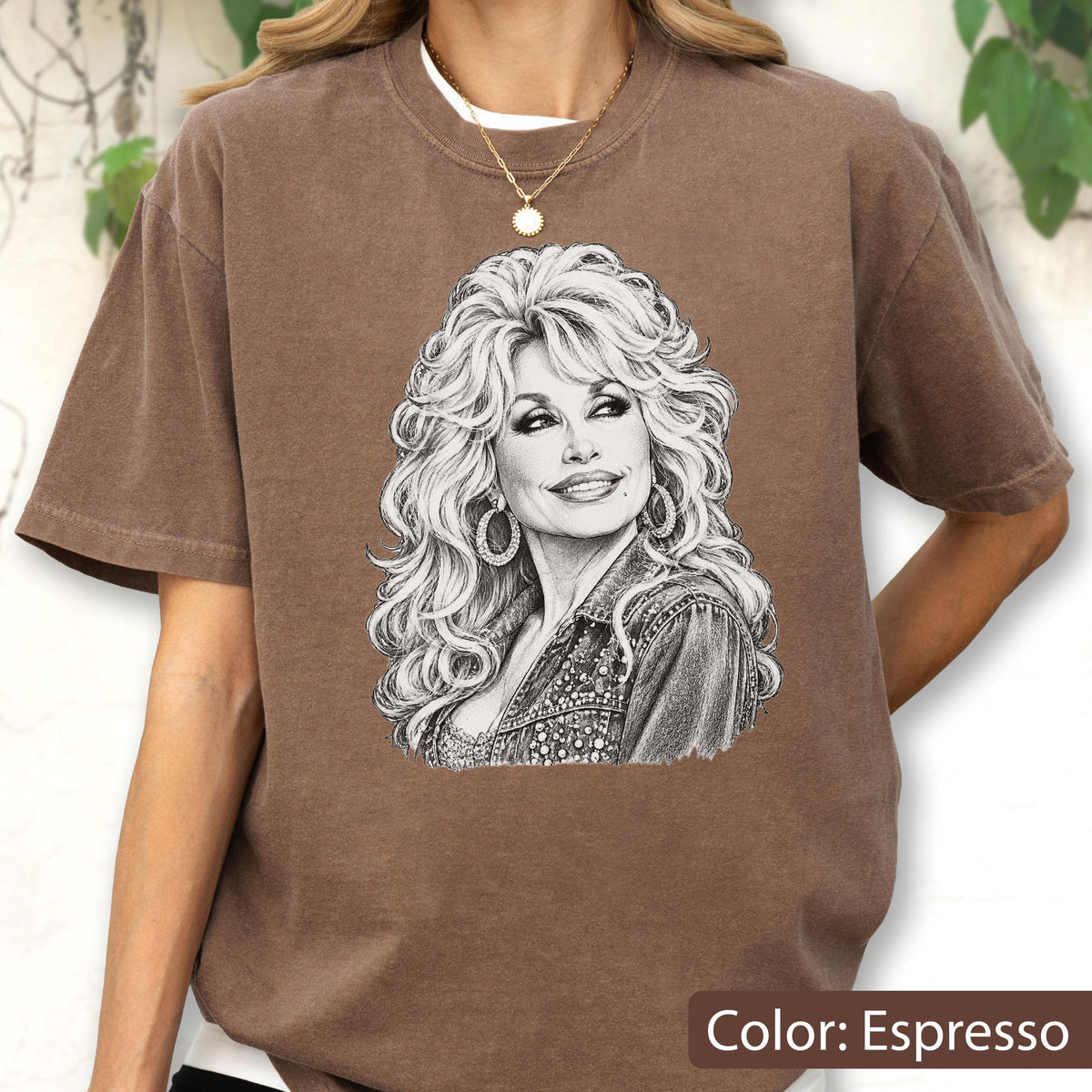 May include: An espresso brown short-sleeve t-shirt featuring a detailed black and white sketch illustration of a woman with voluminous blonde hair, large hoop earrings, and a denim jacket, centered on the front of the garment.