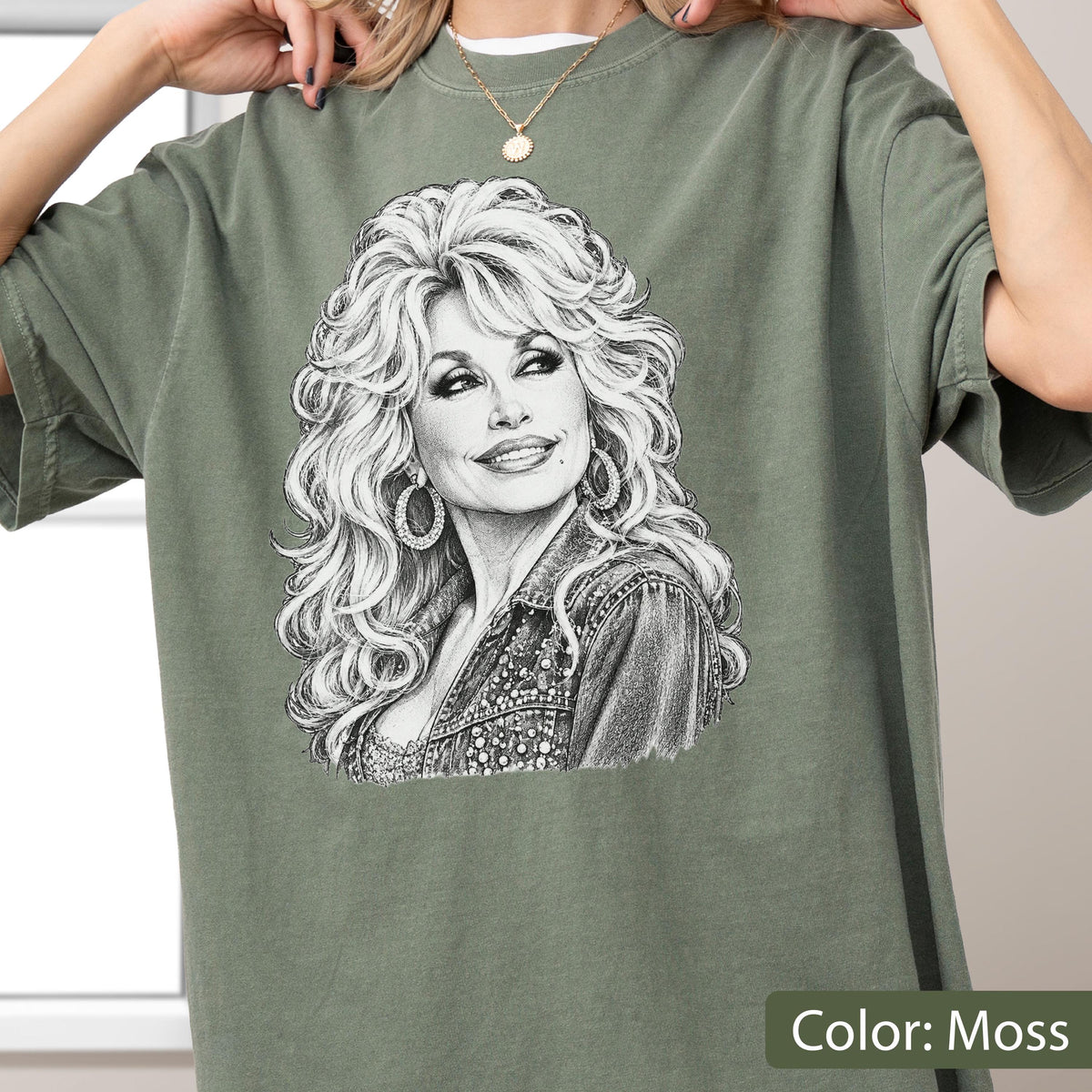 May include: A moss green short-sleeve t-shirt featuring a detailed black and white pencil-style sketch graphic of a person with voluminous blonde hair, wearing hoop earrings and a denim jacket, centered on the front of the garment.