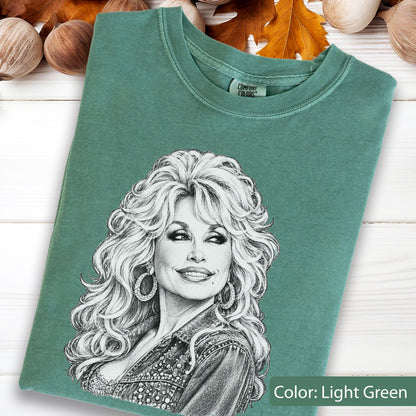 May include: A light green t-shirt featuring a detailed black and white pencil-style sketch of a person with voluminous wavy hair and hoop earrings. The shirt has a crew neckline and a Comfort Colors brand label printed on the inside collar area.