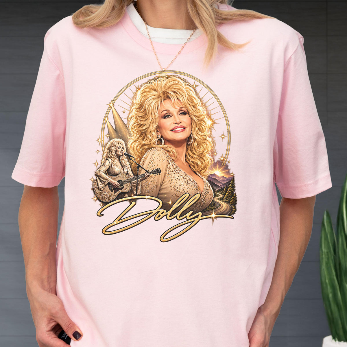 May include: A light pink short-sleeve t-shirt featuring a graphic print of Dolly Parton with blonde hair, a smaller image of her playing an acoustic guitar, a mountain landscape, and the word Dolly in gold cursive script with star accents.