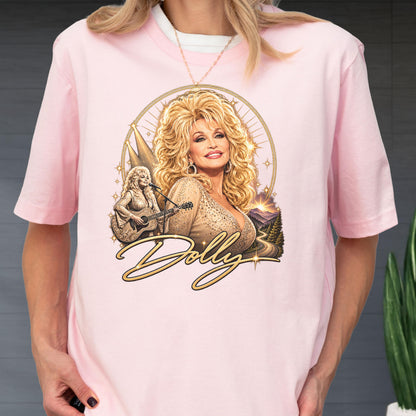 May include: A light pink short-sleeve t-shirt featuring a graphic print of Dolly Parton with blonde hair, a smaller image of her playing an acoustic guitar, a mountain landscape, and the word Dolly in gold cursive script with star accents.