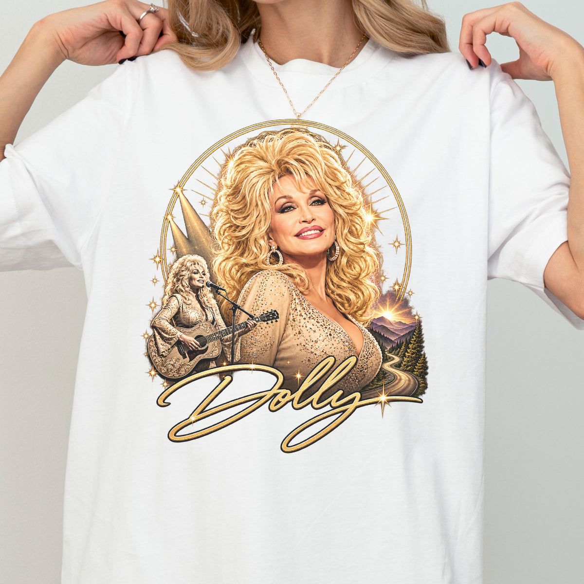 May include: A white t-shirt featuring a graphic print of Dolly Parton with voluminous blonde hair. The design includes a portrait, a smaller image of her playing an acoustic guitar, a mountain landscape, and the script text Dolly in gold.