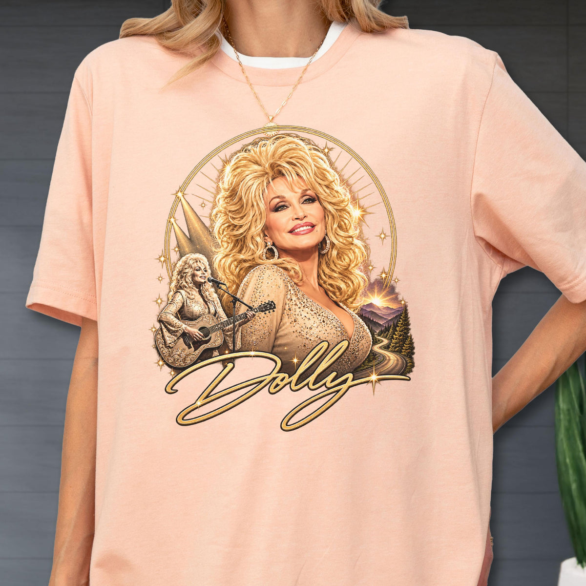May include: A peach-colored t-shirt featuring a graphic print of Dolly Parton with blonde hair, wearing a tan top. The design includes a smaller image of her playing a guitar, a mountain landscape, star accents, and the word Dolly in gold script.