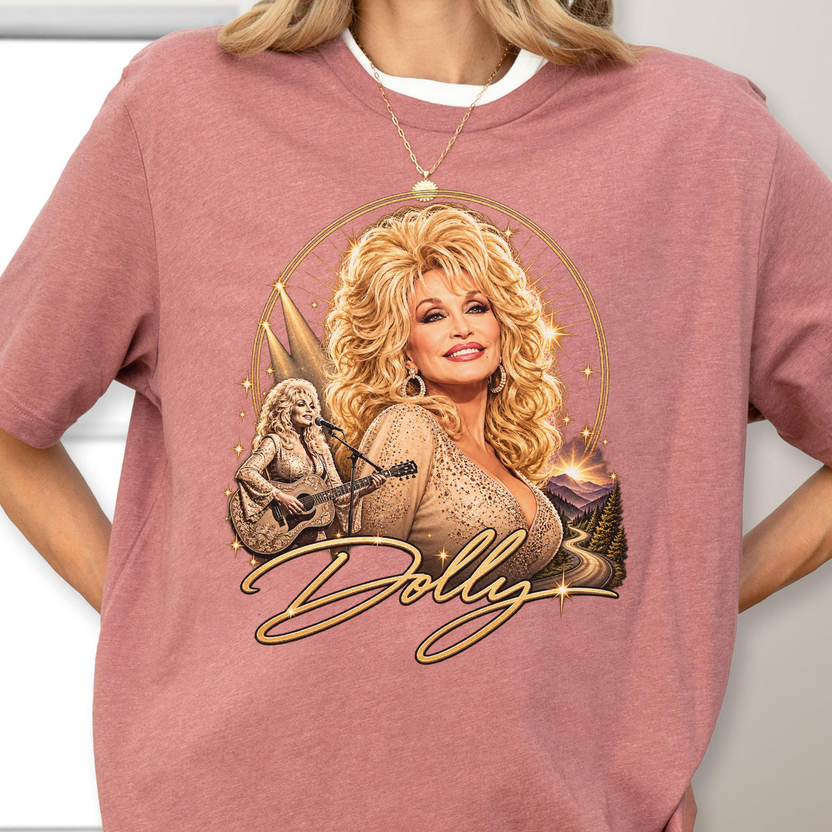 May include: A mauve short-sleeved t-shirt featuring a graphic print of Dolly Parton with blonde hair, wearing a tan top, and playing an acoustic guitar. The design includes a circular frame, stars, a mountain landscape, and the name Dolly in script.