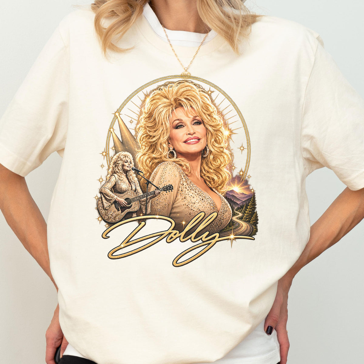 May include: A cream-colored t-shirt featuring a graphic print of a blonde woman with voluminous hair, wearing a shimmering top. The design includes a smaller image of the woman playing an acoustic guitar, a mountain landscape, and the word Dolly.