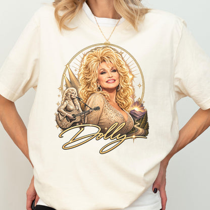 May include: A cream-colored t-shirt featuring a graphic print of a blonde woman with voluminous hair, wearing a shimmering top. The design includes a smaller image of the woman playing an acoustic guitar, a mountain landscape, and the word Dolly.