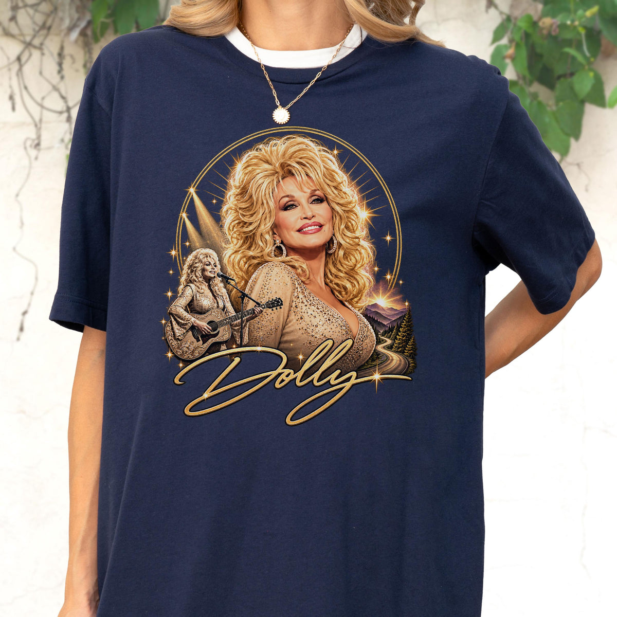 May include: A navy blue short-sleeve t-shirt featuring a graphic print of Dolly Parton with blonde hair, wearing a tan top. The design includes a smaller image of her playing an acoustic guitar, a mountain landscape, and the script text Dolly.