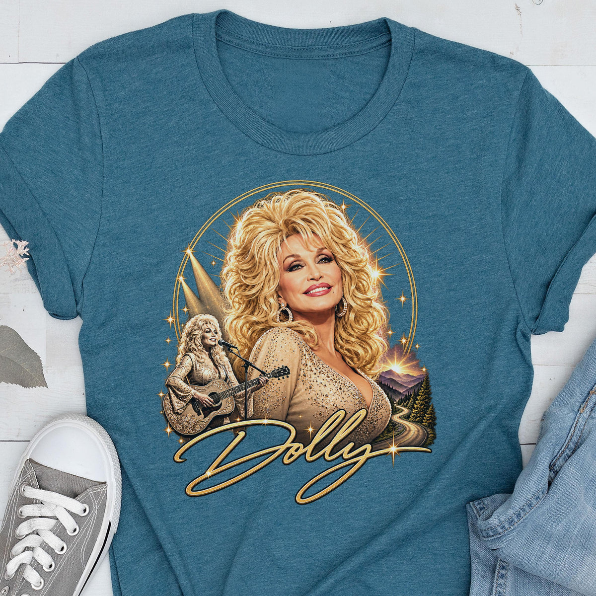 May include: A teal crew neck t-shirt featuring a graphic print of a blonde woman with voluminous hair. The design includes a smaller image of the woman playing an acoustic guitar, a golden circular frame, and the cursive text Dolly in gold.
