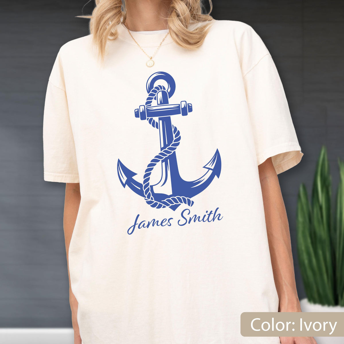 May include: An ivory short-sleeve t-shirt featuring a large blue graphic of a nautical anchor wrapped in a rope. Below the anchor, the text James Smith is printed in a blue script font. The shirt has a crew neckline and a relaxed fit.