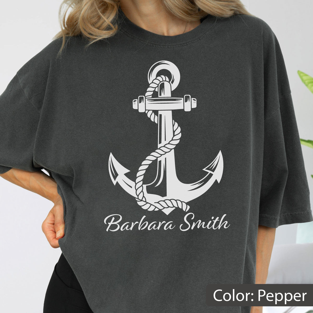 May include: A charcoal gray short-sleeve t-shirt featuring a large white graphic of a nautical anchor wrapped in a thick rope. Below the anchor, the name Barbara Smith is printed in a white cursive script font. The shirt has a crew neckline.