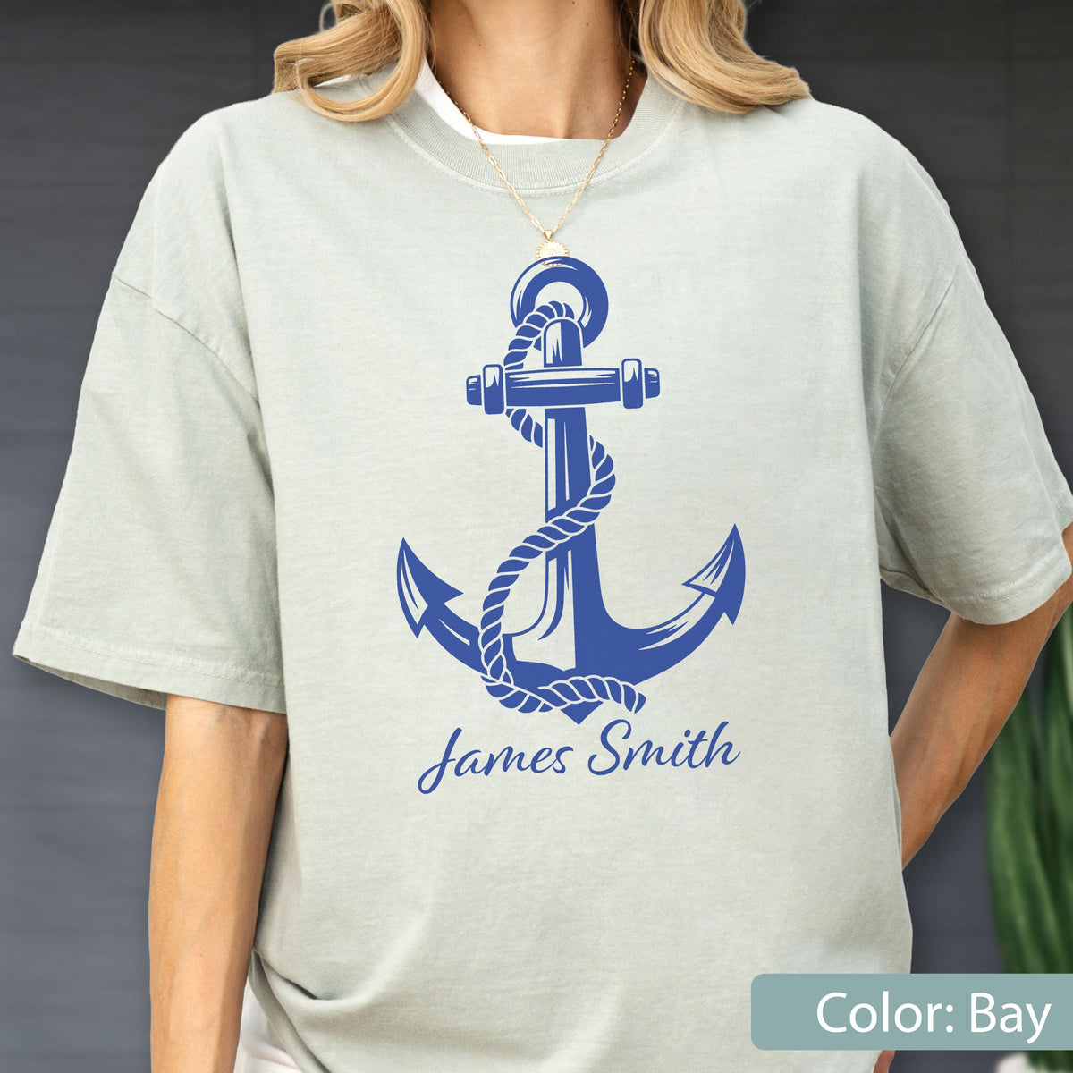 May include: A light sage green short-sleeve t-shirt featuring a large blue graphic of a nautical anchor wrapped in rope. Below the anchor, the name James Smith is printed in a blue script font. The shirt has a crew neckline and a relaxed fit.