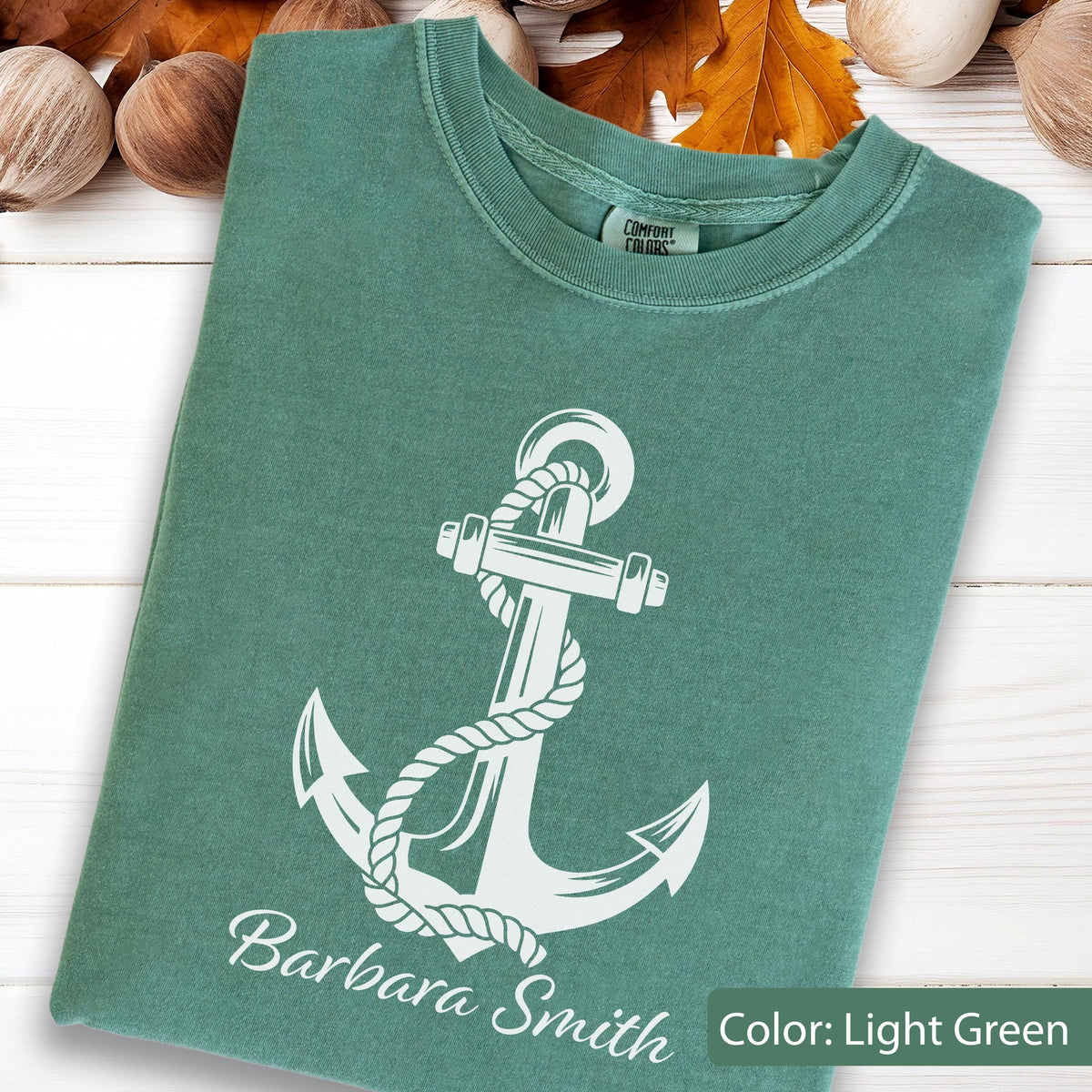 May include: A light green crew neck t-shirt featuring a white graphic of a nautical anchor wrapped in rope. The text "Barbara Smith" is printed in white below the anchor. The shirt has a Comfort Colors brand label inside the neckline.