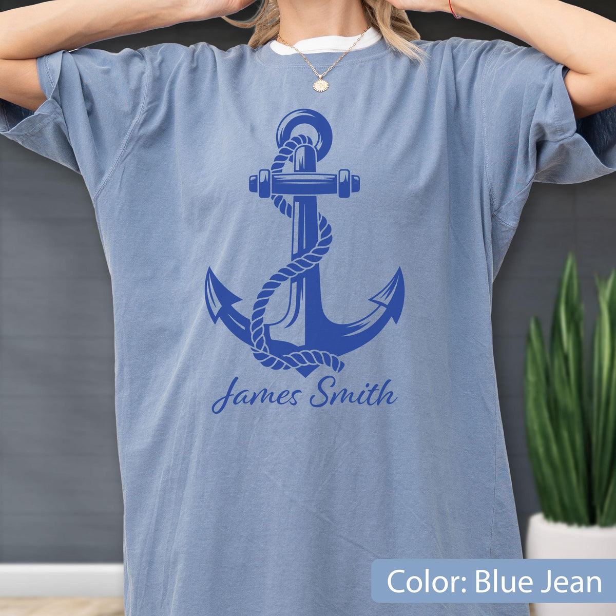May include: A blue jean colored t-shirt featuring a large, dark blue graphic of a ship anchor wrapped in rope. Below the anchor, the name James Smith is printed in a dark blue cursive script font. The shirt has short sleeves and a crew neckline.