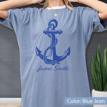 May include: A blue jean colored t-shirt featuring a large, dark blue graphic of a ship anchor wrapped in rope. Below the anchor, the name James Smith is printed in a dark blue cursive script font. The shirt has short sleeves and a crew neckline.