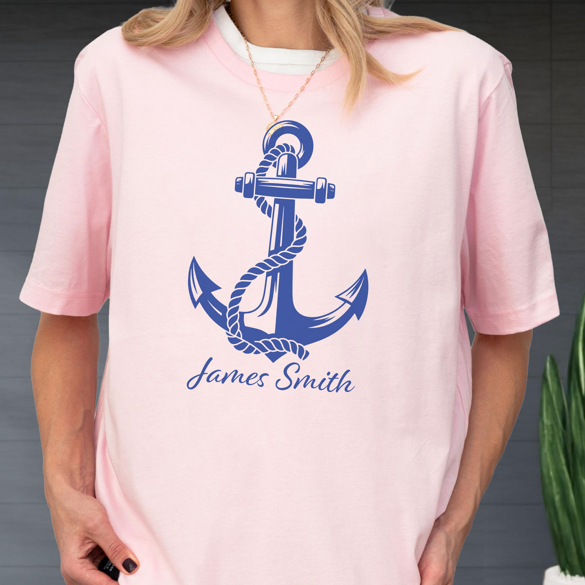 May include: A light pink short-sleeved t-shirt featuring a large dark blue graphic of a nautical anchor wrapped in a rope. Below the anchor, the name James Smith is printed in a dark blue cursive script font on the front of the garment.