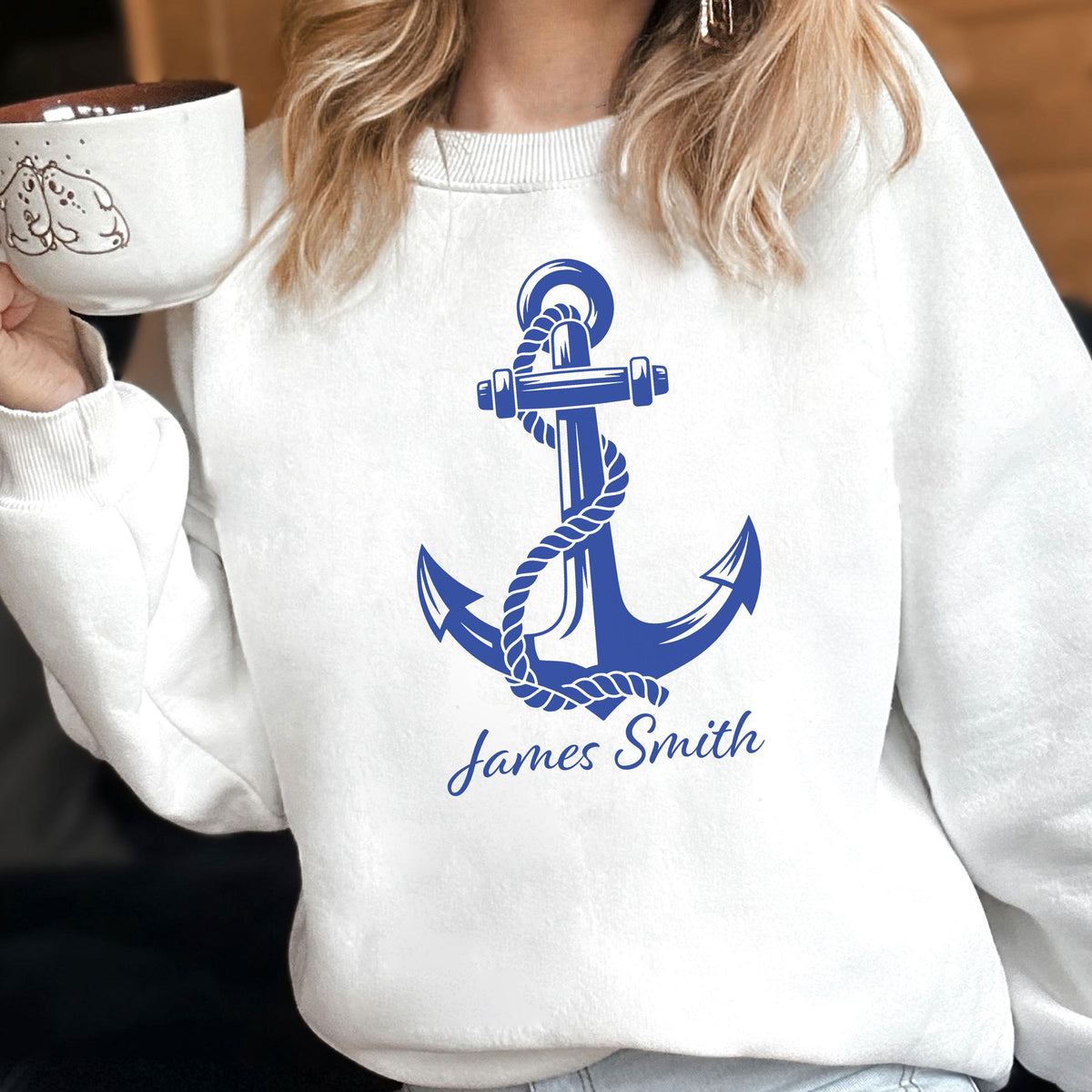 May include: A white crewneck sweatshirt featuring a large blue graphic of a nautical anchor wrapped in a rope. Below the anchor, the text James Smith is printed in a blue script font. The sweatshirt has long sleeves and a ribbed neckline.