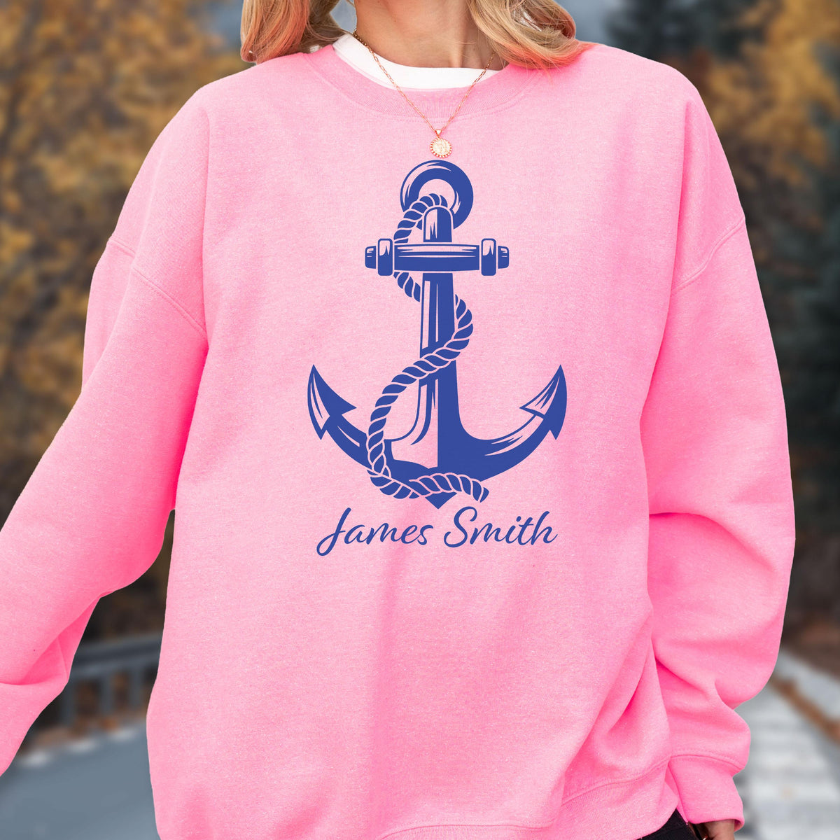 May include: A bright pink crewneck sweatshirt featuring a large, dark blue graphic of a nautical anchor wrapped in rope. Below the anchor, the name James Smith is printed in a dark blue script font. The garment has long sleeves and a ribbed collar.