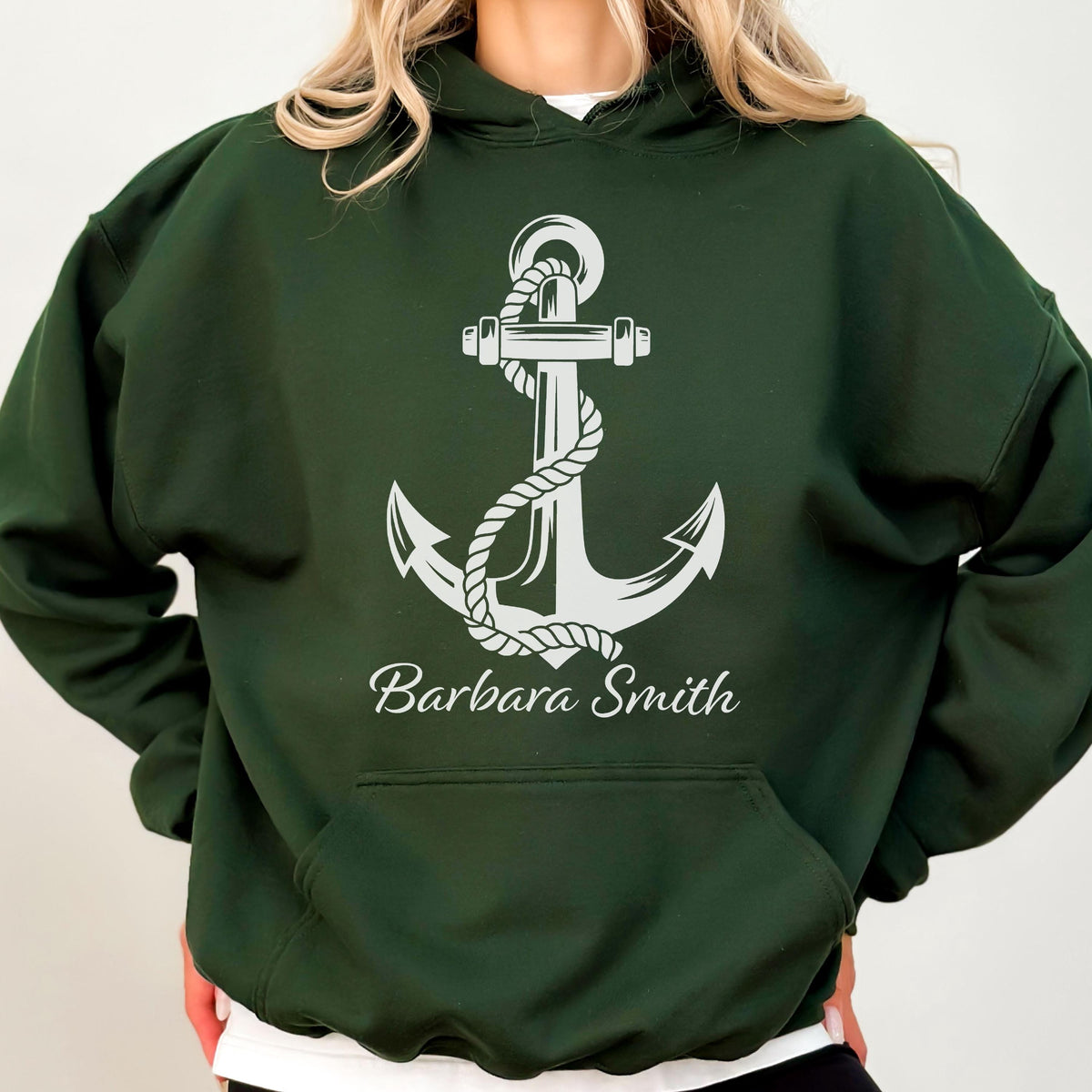 May include: A dark forest green hooded sweatshirt featuring a large white graphic of a nautical anchor wrapped in a rope. Below the anchor, the name Barbara Smith is printed in a white cursive script font. The hoodie includes a front kangaroo pocket.