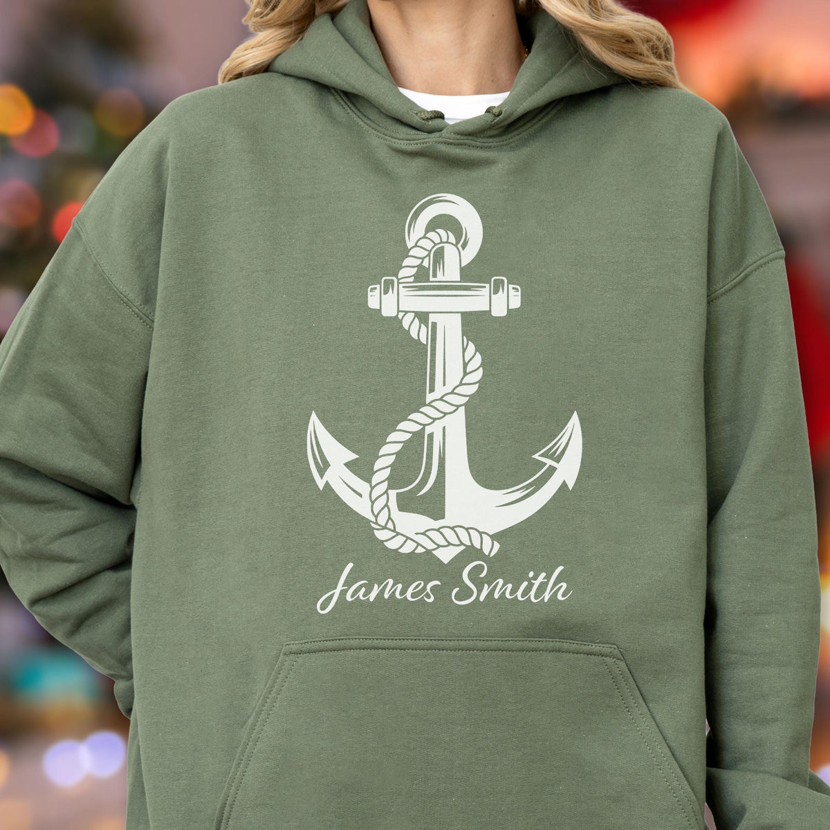 May include: An olive green hooded sweatshirt featuring a large white graphic of a nautical anchor wrapped in rope. Below the anchor, the name James Smith is printed in a white script font. The garment includes a front kangaroo pocket.