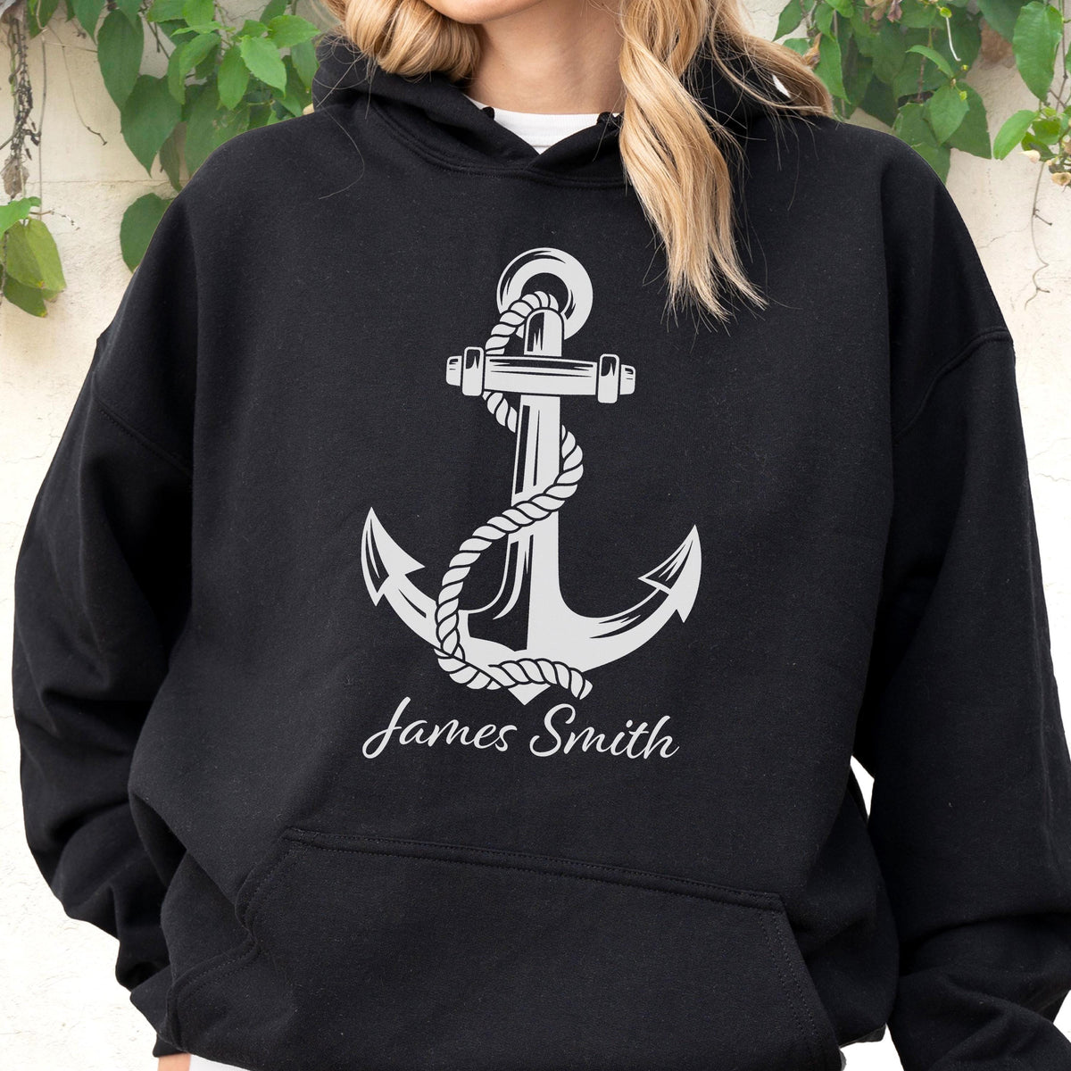 May include: A black hooded sweatshirt featuring a large white graphic of a nautical anchor wrapped in rope. Below the anchor, the name James Smith is printed in a white script font. The garment includes a front kangaroo pocket and long sleeves.