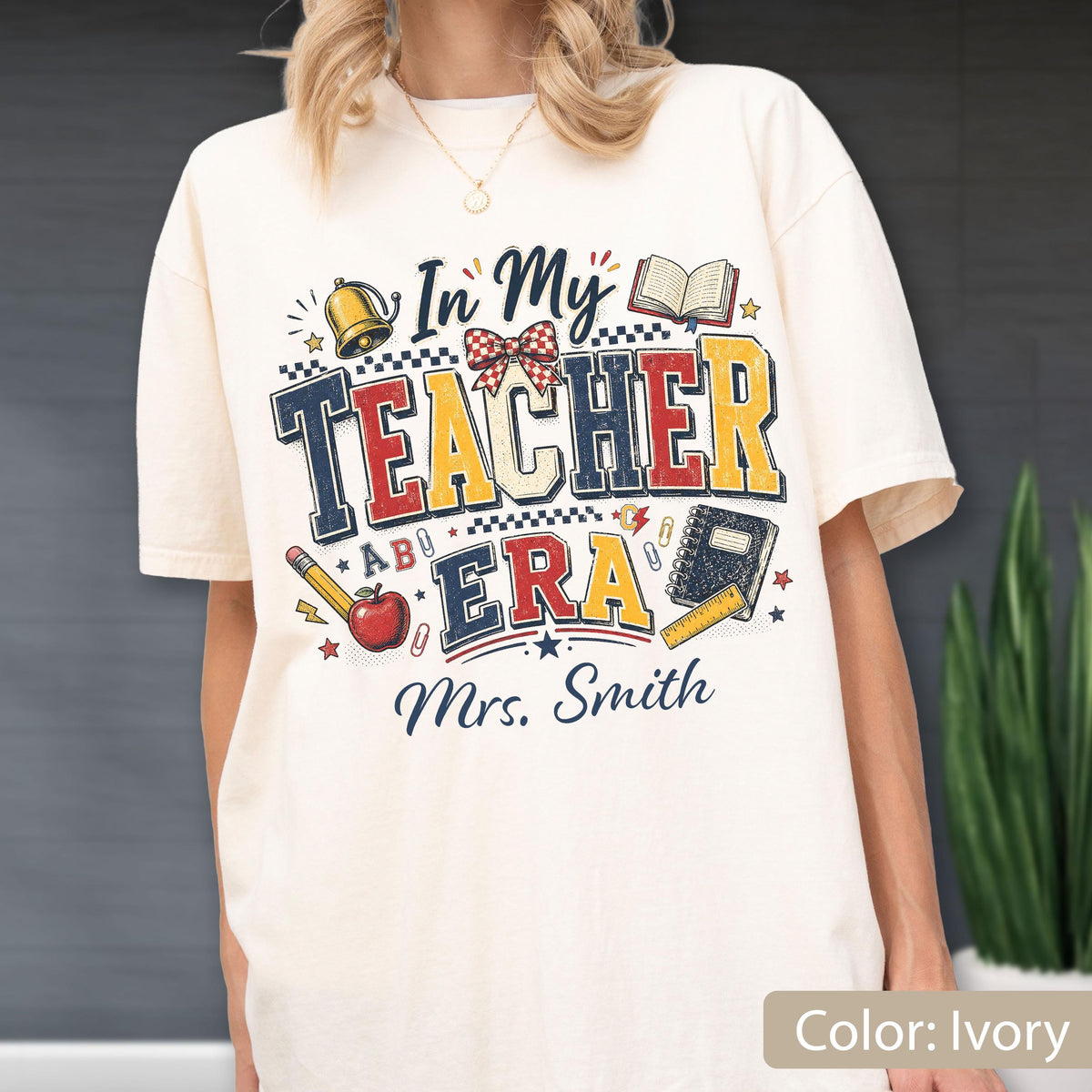 May include: An ivory t-shirt featuring a graphic print with the text "In My TEACHER ERA" and "Mrs. Smith". The design includes illustrations of a gold bell, an open book, a red apple, a pencil, a notebook, a ruler, and a red and white bow.