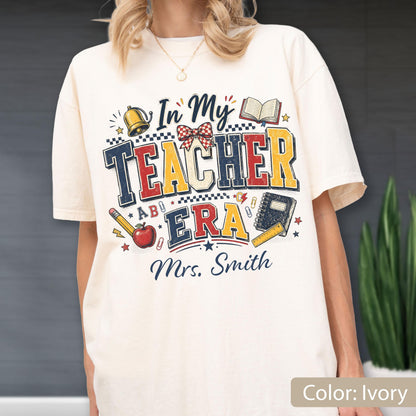 May include: An ivory t-shirt featuring a graphic print with the text "In My TEACHER ERA" and "Mrs. Smith". The design includes illustrations of a gold bell, an open book, a red apple, a pencil, a notebook, a ruler, and a red and white bow.