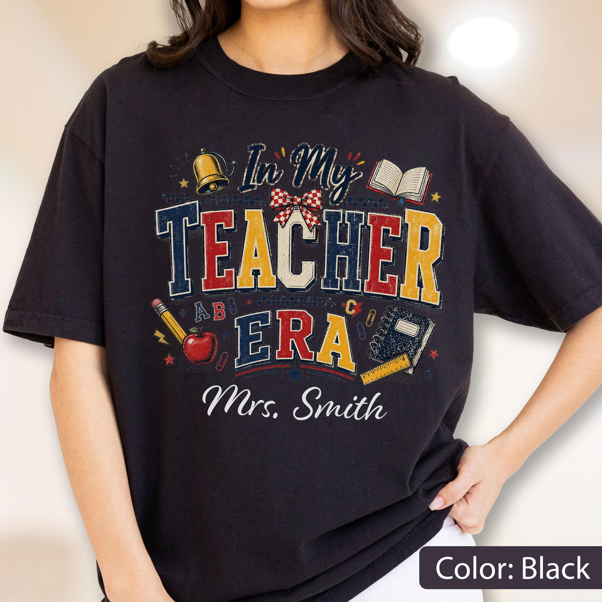 May include: A black t-shirt featuring a graphic with the text In My Teacher Era and Mrs. Smith. The design includes a gold bell, a red apple, a pencil, an open book, a notebook, a ruler, and a red and white checkered bow in a distressed style.