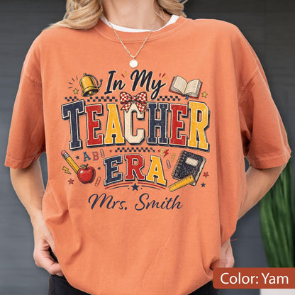 May include: An orange short-sleeve t-shirt featuring a graphic print with the text "In My TEACHER ERA" and "Mrs. Smith". The design includes illustrations of a bell, an open book, a pencil, an apple, a notebook, and a ruler with stars and patterns.