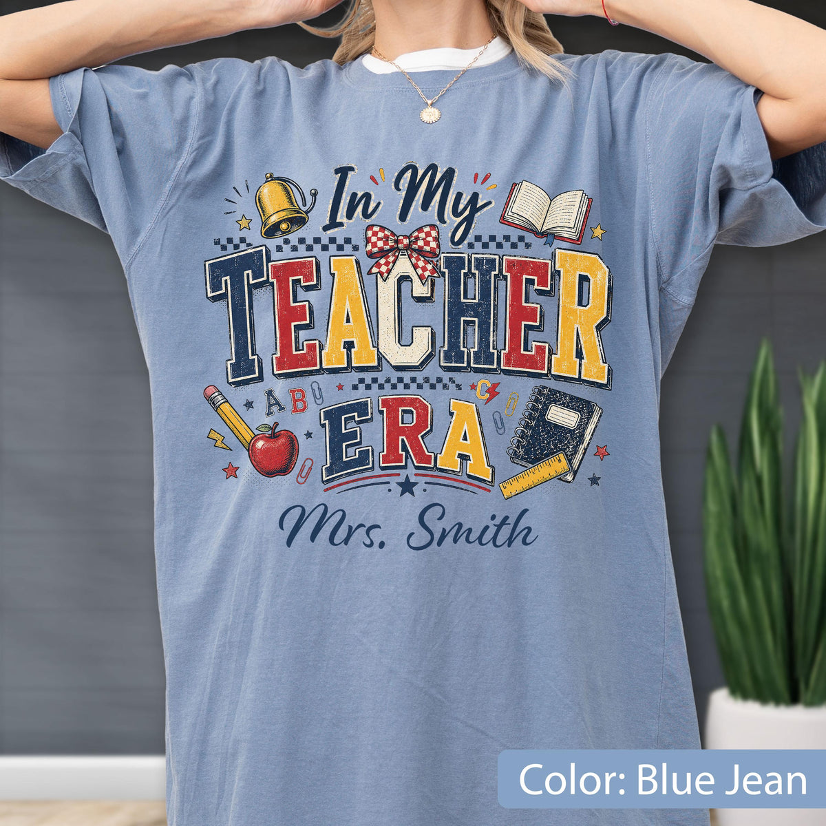 May include: A blue jean colored t-shirt featuring the text In My Teacher Era and Mrs. Smith. The graphic includes a gold bell, an open book, a red apple, a pencil, a notebook, a ruler, and a red and white checkered bow in a collegiate style.