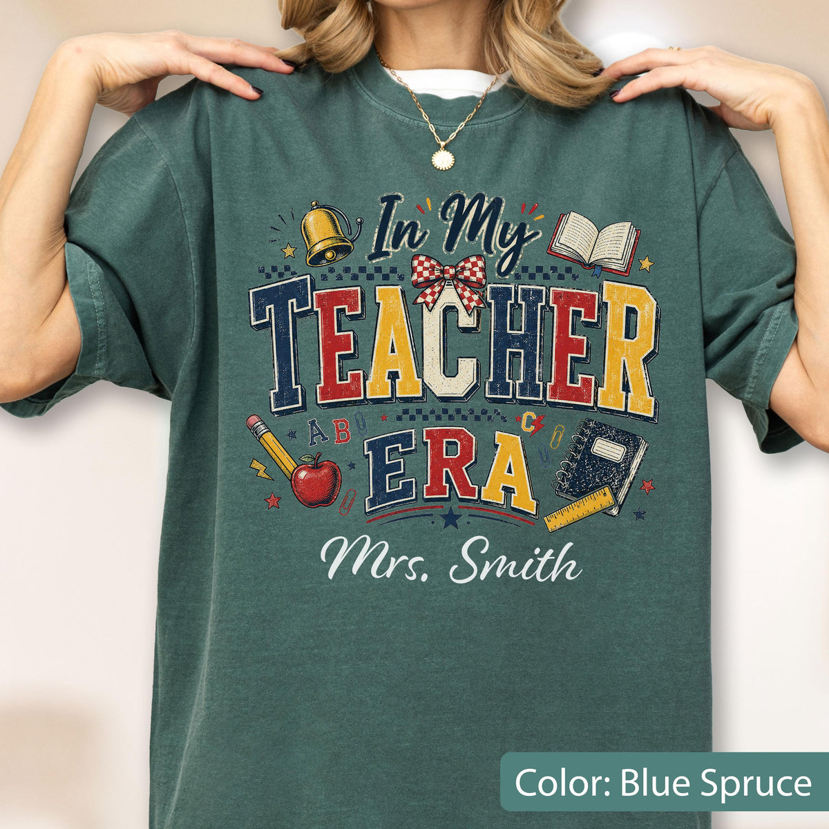 May include: A blue-spruce t-shirt featuring a graphic with the text In My Teacher Era and Mrs. Smith. The design includes a gold bell, a red and white checkered bow, an open book, a pencil, a red apple, a notebook, and a yellow ruler.