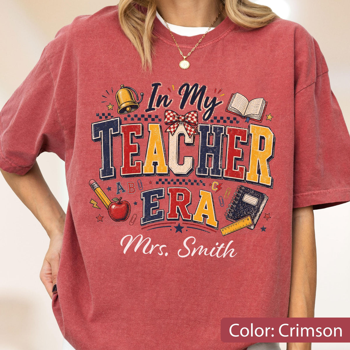 May include: A crimson t-shirt featuring a graphic with the text In My Teacher Era and Mrs. Smith. The design includes a gold bell, a red apple, a pencil, an open book, a notebook, a ruler, and a red and white checkered bow on a dark background.