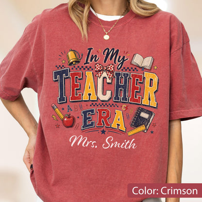 May include: A crimson t-shirt featuring a graphic with the text In My Teacher Era and Mrs. Smith. The design includes a gold bell, a red apple, a pencil, an open book, a notebook, a ruler, and a red and white checkered bow on a dark background.