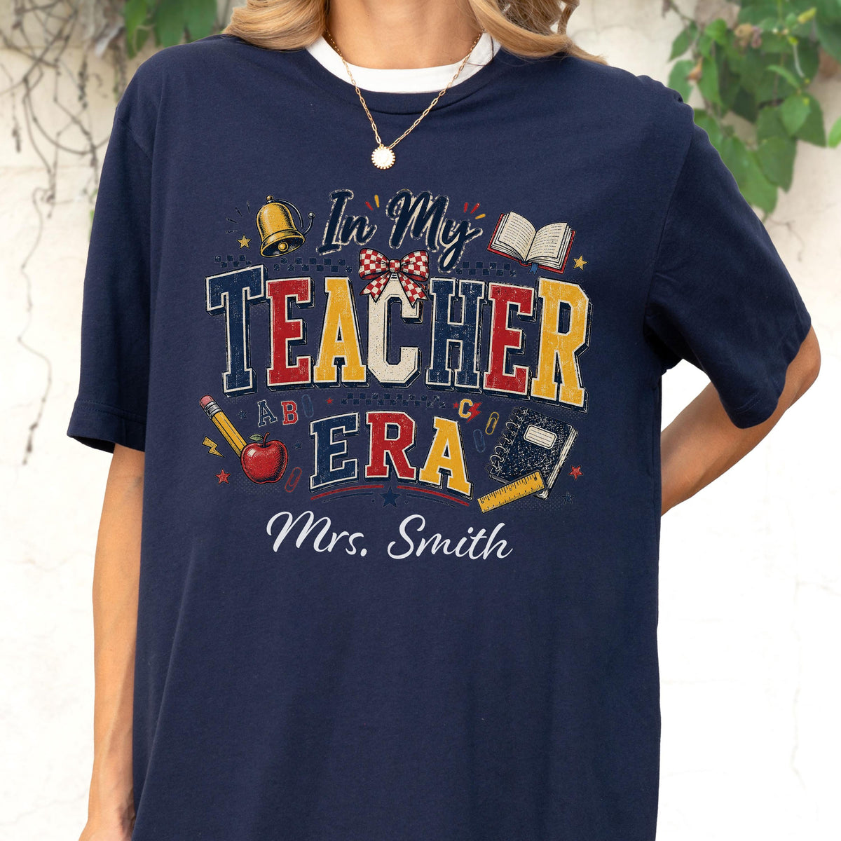 May include: A navy blue t-shirt featuring a graphic print with the text "In My TEACHER ERA" and "Mrs. Smith". The design includes a gold bell, a red and white checkered bow, an open book, a yellow pencil, a red apple, and a black notebook.