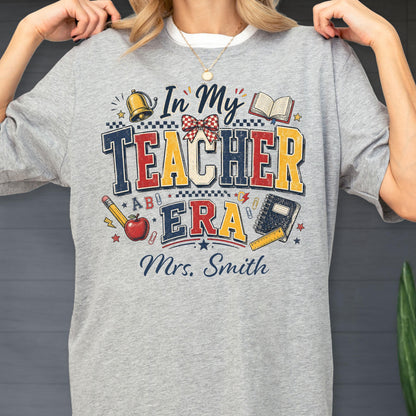 May include: A heather gray t-shirt featuring the text In My Teacher Era and Mrs. Smith in navy blue. The design includes a gold bell, a red and white checkered bow, an open book, a yellow pencil, a red apple, a black notebook, and a yellow ruler.