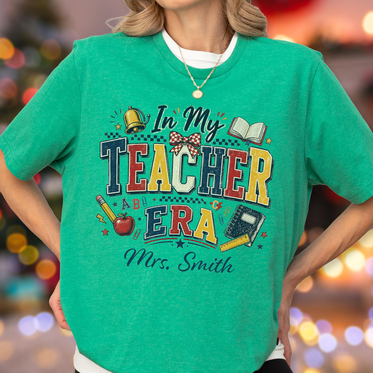 May include: A bright green short-sleeve t-shirt featuring a graphic print with the text In My Teacher Era and Mrs. Smith. The design includes illustrations of a bell, a bow, an open book, a pencil, an apple, and a notebook with a ruler.