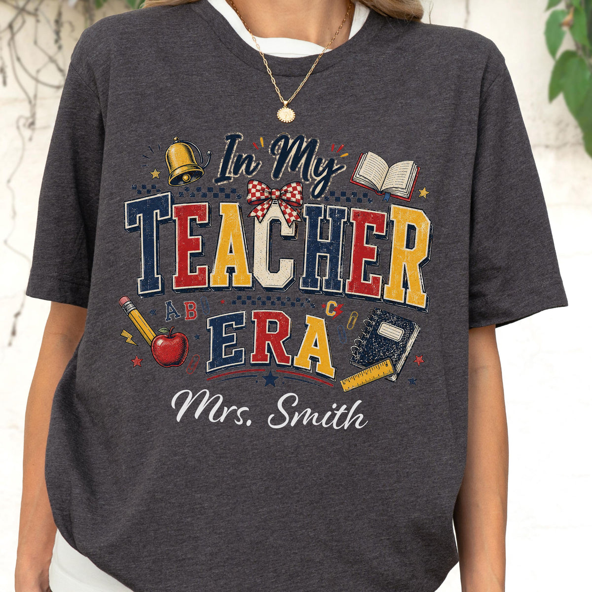 May include: A dark heather gray t-shirt featuring a graphic print with the text In My Teacher Era and Mrs. Smith. The design includes a gold bell, an open book, a red apple, a pencil, a notebook, and a red and white checkered bow.