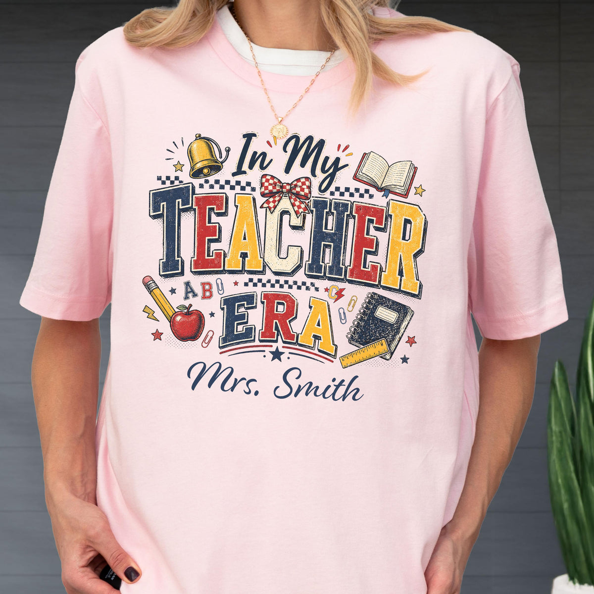 May include: A light pink t-shirt featuring a graphic print with the text In My Teacher Era and Mrs. Smith. The design includes illustrations of a bell, an open book, a red apple, a pencil, a notebook, a ruler, and a checkered bow in red and white.