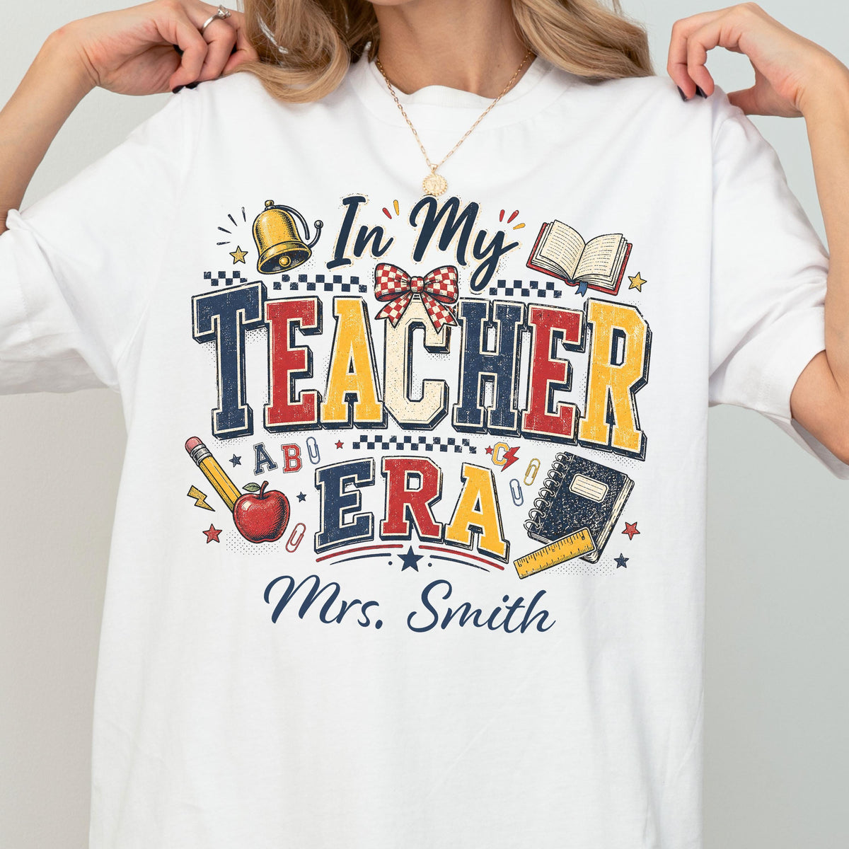 May include: A white t-shirt featuring a graphic design with the text In My Teacher Era and Mrs. Smith. The design includes a gold bell, an open book, a red apple, a pencil, a spiral notebook, a ruler, a red and white checkered bow, and paper clips.