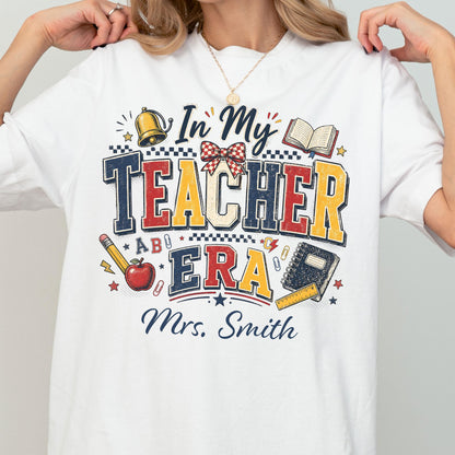 May include: A white t-shirt featuring a graphic design with the text In My Teacher Era and Mrs. Smith. The design includes a gold bell, an open book, a red apple, a pencil, a spiral notebook, a ruler, a red and white checkered bow, and paper clips.