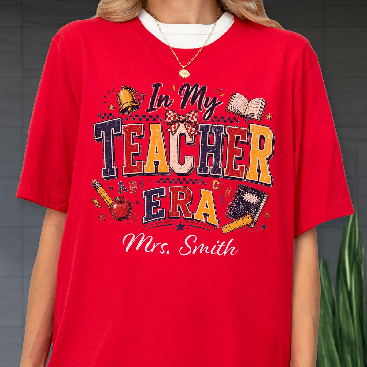 May include: A bright red short-sleeved t-shirt featuring a graphic print with the text In My Teacher Era and Mrs. Smith. The design includes illustrations of a gold bell, a red apple, a pencil, a notebook, and a ruler in various colors.