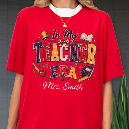 May include: A bright red short-sleeved t-shirt featuring a graphic print with the text In My Teacher Era and Mrs. Smith. The design includes illustrations of a gold bell, a red apple, a pencil, a notebook, and a ruler in various colors.