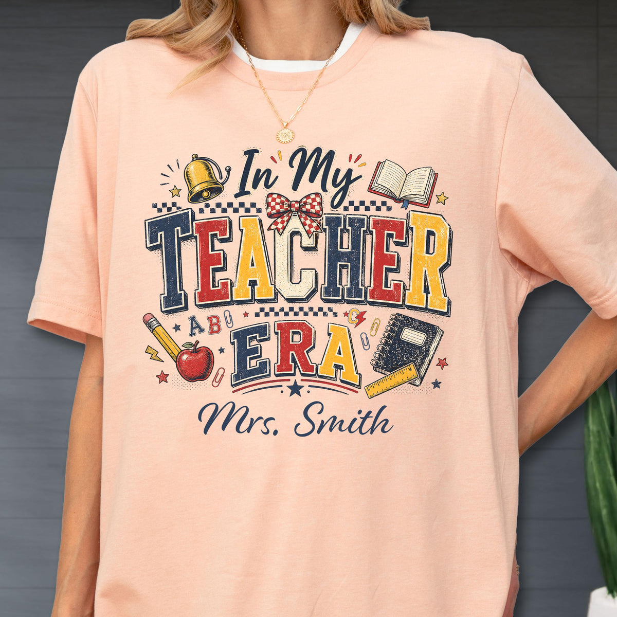 May include: A peach-colored t-shirt featuring a graphic with the text In My Teacher Era and Mrs. Smith. The design includes a gold bell, an open book, a red apple, a pencil, a notebook, a ruler, and a red and white checkered bow in a vintage style.