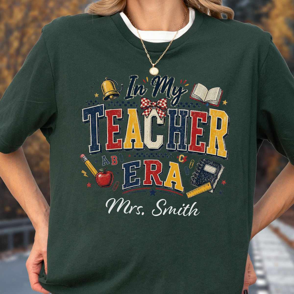 May include: A dark green t-shirt featuring a graphic with the text In My Teacher Era and Mrs. Smith. The design includes a gold bell, a red apple, a pencil, a ruler, a notebook, an open book, and a red and white checkered bow in a collegiate font.