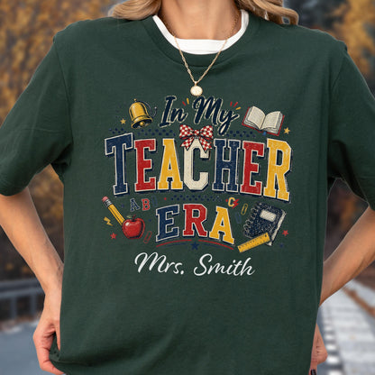 May include: A dark green t-shirt featuring a graphic with the text In My Teacher Era and Mrs. Smith. The design includes a gold bell, a red apple, a pencil, a ruler, a notebook, an open book, and a red and white checkered bow in a collegiate font.